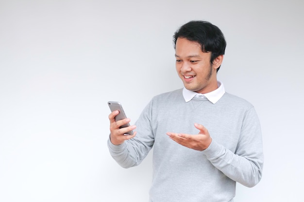 若いアジア人男性は、孤立した灰色の背景のスマートフォンで彼が見るものにショックを受けて満足しています