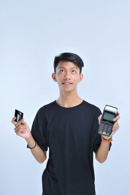 Молодой азиатский мужчина показывает кредитную / дебетовую карту и машину электронного сбора данных (EDC) для простой и быстрой финансовой транзакции, изолированной на сером фоне