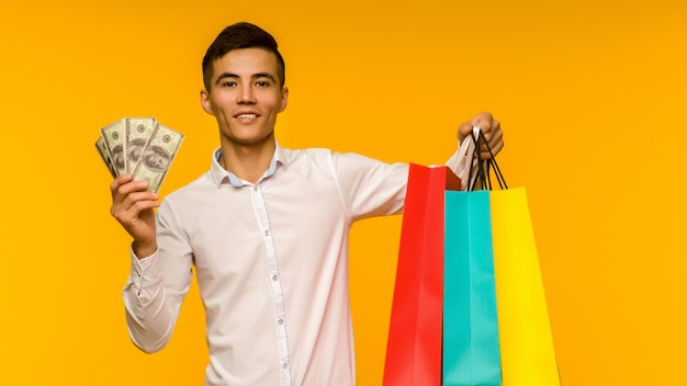 노란색 배경에 그의 쇼핑백과 돈을 보여주는 젊은 아시아 남자