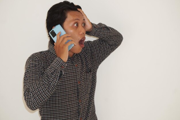 Photo young asian man showing shocked face expression while in call using handphone device