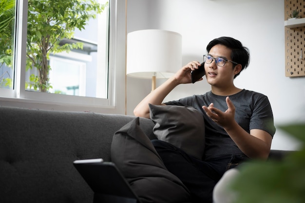 自宅のソファに座って携帯電話で話している若いアジア人男性。