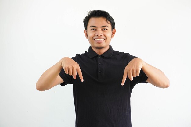 Photo young asian man smiling at the camera and pointing below wearing black polo t shirt isolated