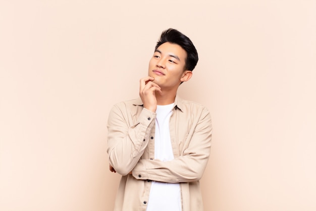 Young asian man smiling happily and daydreaming or doubting, looking to the side over color wall