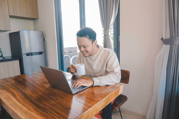 A young Asian man spends his time at home sitting in the dining room happily working on his laptop while holding a mug