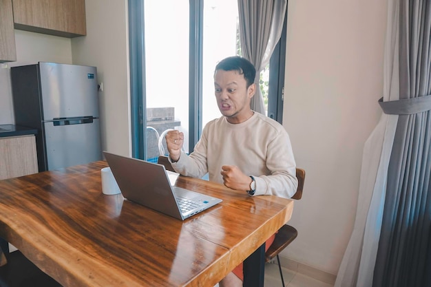 A young Asian man spends his time at home sitting in the dining room working on his laptop feel frustrated