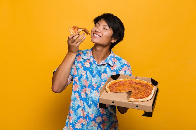 Young asian man standing isolated over yellow space eat pizza.