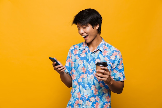 Young asian man standing isolated over yellow space using mobile phone drinking coffee.