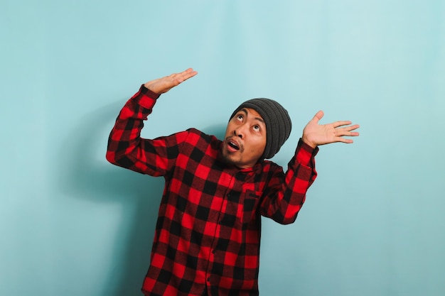 Young Asian man stands cowering avoiding danger above him while standing against a blue background