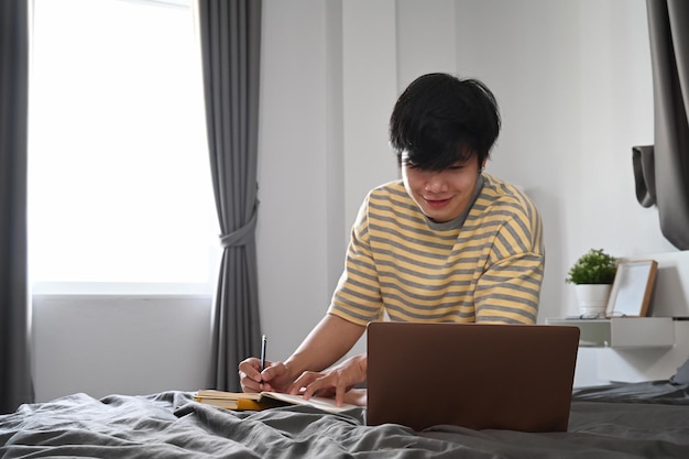 Young asian man studying online with laptop computer in bedroom.