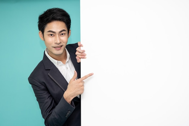 Young asian man in suit standing behind blank white billboard and pointing to empty copy space