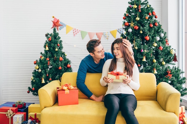 Young Asian man surprises her girlfriend that wearing Santa Claus hat with a Christmas gift at home with a Christmas tree in the background Image with copy space