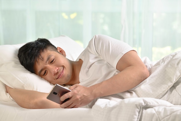 Young Asian man using smartphone in bed after waking up in the morning