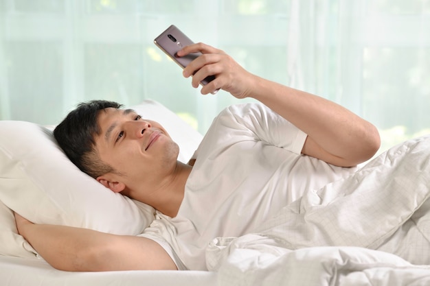 Young Asian man using smartphone in bed after waking up in the morning