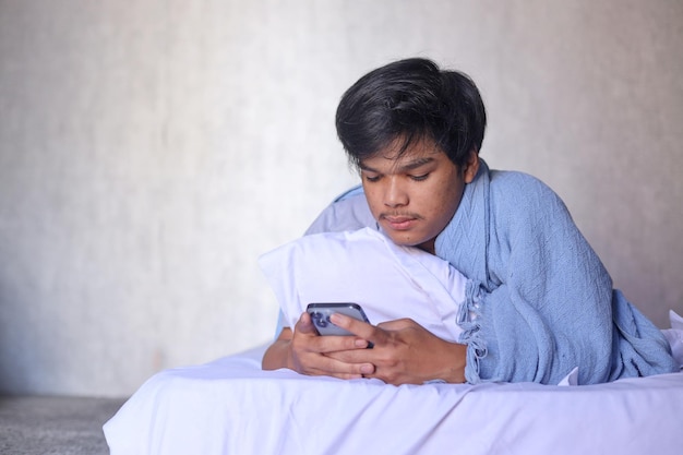 Young Asian man using smartphone in bed after waking up in the morning