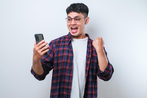 Young asian man using smartphone screaming proud and celebrating victory and success very excited cheering emotion