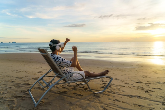 Giovane uomo asiatico che utilizza occhiali per realtà virtuale per riunioni di lavoro sulla spiaggia tropicale su uno sfondo bellissimo mare e cielo