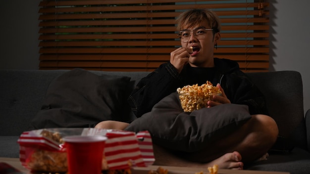 Giovane uomo asiatico che guarda la tv e mangia popcorn sul divano godendo di attività ricreative a casa