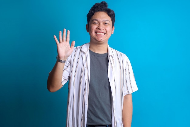 Young Asian man waving hand saying hello goodbye on blue background