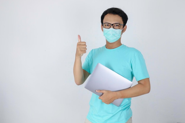 Young Asian Man wear medical mask is carrying a laptop with ok hand sign Indonesian man wearing blue shirt