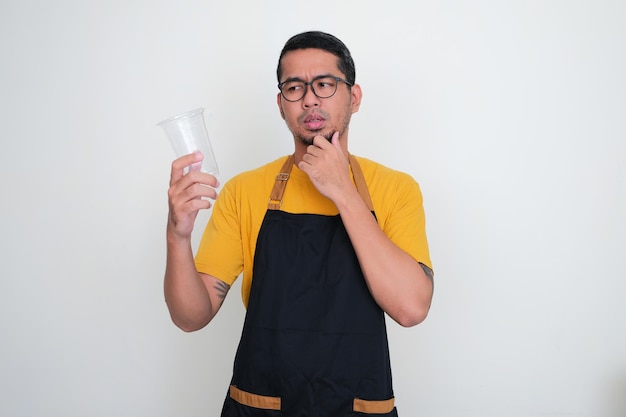 エプロンを着た若いアジア人男性が、手に持ったプラスチックのコップを見ながら考える表情を見せる