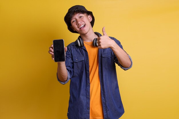 Young Asian man wearing bucket hat and headphones is smiling and give thumb up while showing blank m