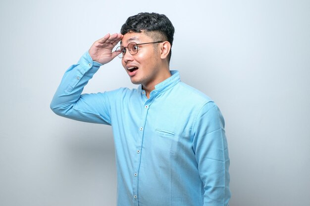 Il giovane uomo asiatico che indossa una camicia casual e occhiali sembra molto felice e sorridente guardando lontano con la mano sopra la testa