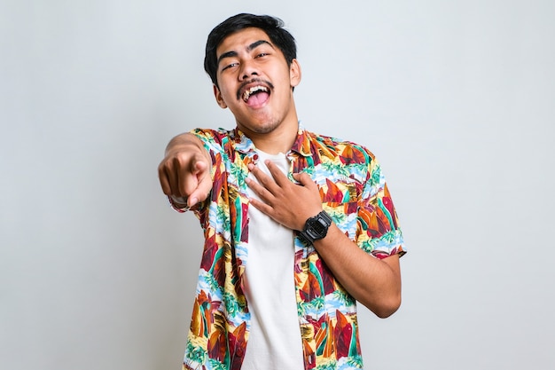 Young asian man wearing casual shirt laughing at you, pointing finger to the camera with hand over body, shame expression