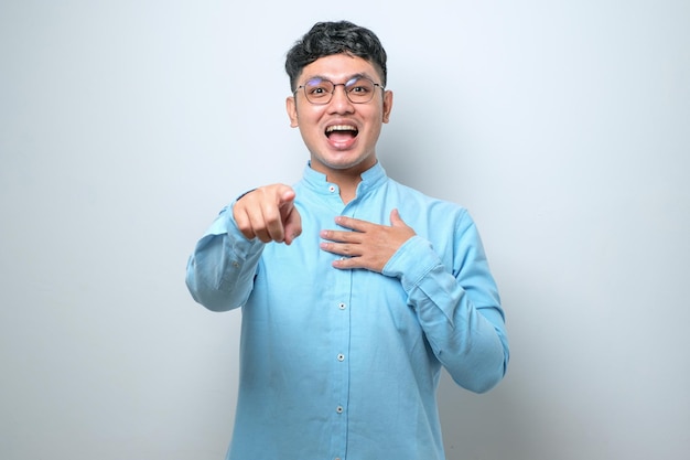 Young asian man wearing casual shirt laughing at you pointing finger to the camera with hand over body shame expression