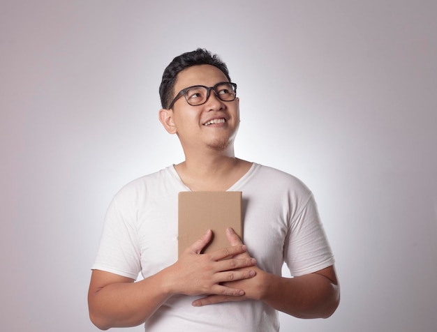 Young Asian man wearing casual white shirt holding a book with thinking gesture and smile