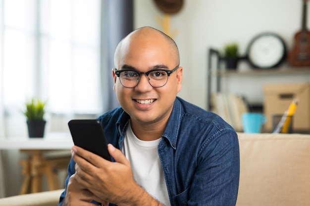 Young asian man wearing glasses sitting on sofa in living room and using smartphone