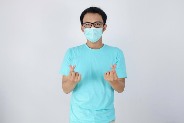 Молодой азиатский мужчина в маске для защиты от болезней показывает знак любви корея на изолированном белом фоне Концепция вируса короны Индонезийский мужчина в синей рубашке