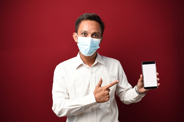 Young asian man wearing medical mask very happy pointing smartphone with blank screen