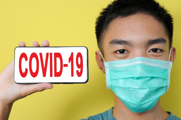 Photo young asian man wearing protect mask showing covid-19 text on phone screen beside his head, isolated on yellow background