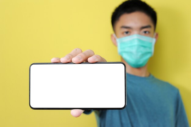 Young Asian man wearing protect mask showing smartphone with blank screen, Isolated on yellow