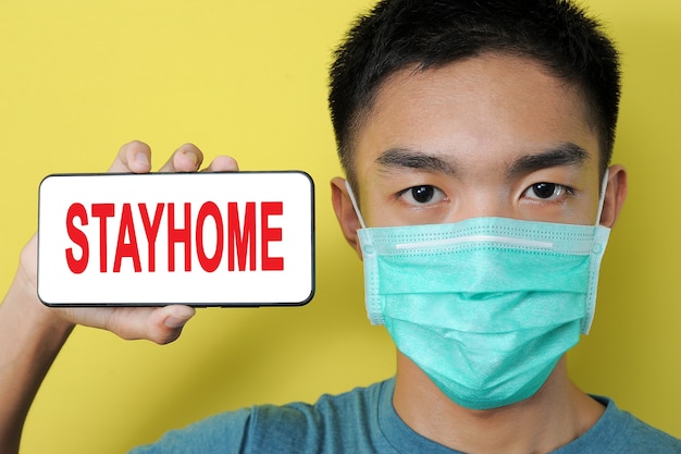 Young Asian man wearing protect mask showing STAYHOME text on phone screen beside his head, isolated on yellow background