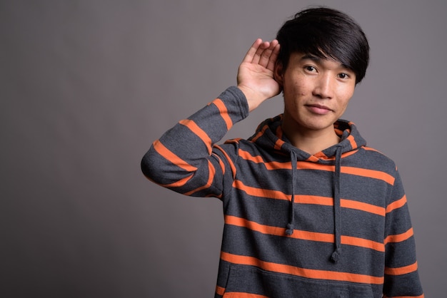Young Asian man wearing striped hoodie against gray wall