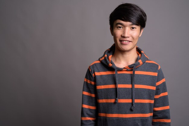 Young Asian man wearing striped hoodie against gray wall