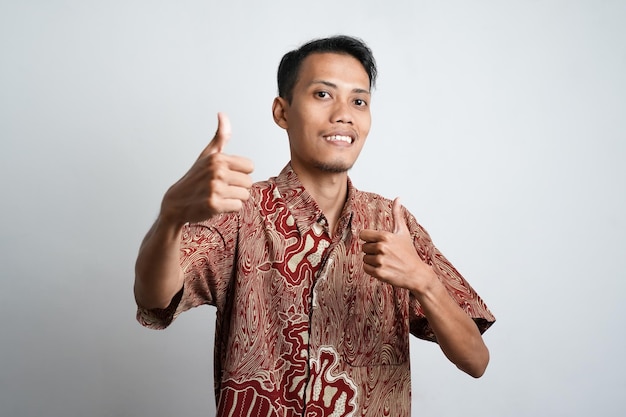 Foto giovane uomo asiatico con la pelle marrone che indossa una camicia batik espressione felice con gioia e mostrando un pollice in su se è d'accordo buon sfondo bianco di prim'ordine