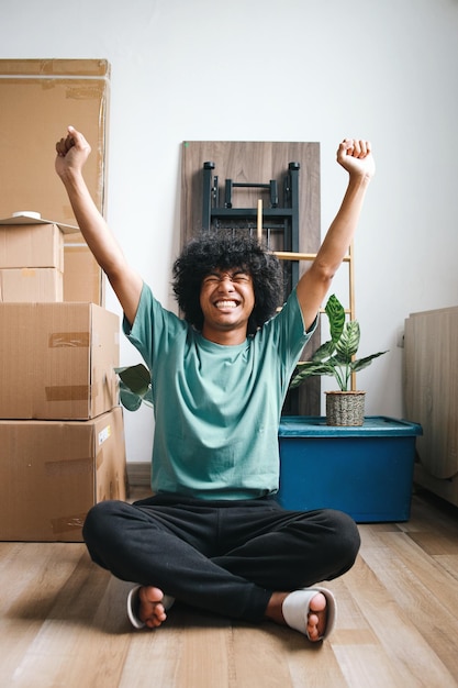 Photo young asian man with curly hair sitting on floor and raising hands celebrating victory after moving