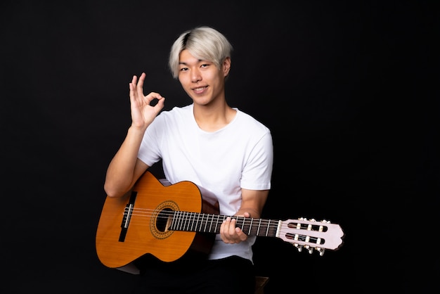 Молодой азиатский человек с гитарой над чернотой показывая одобренный знак с пальцами