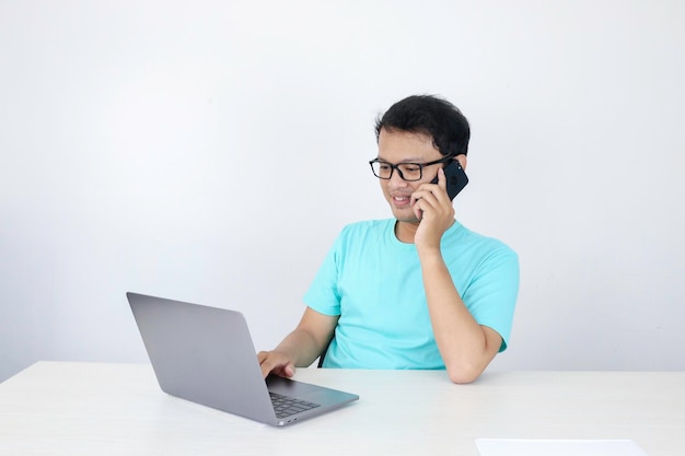 Il giovane uomo asiatico con la faccia felice sta parlando su un telefono cellulare con un laptop sul tavolo uomo indonesiano che indossa una camicia blu