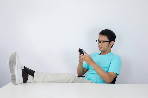 Young Asian Man with happy face with what he see on a mobile phone with leg on the table Indonesian man wearing blue shirt