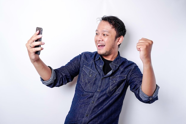 A young Asian man with a happy successful expression wearing blue shirt and holding his phone isolated by white background