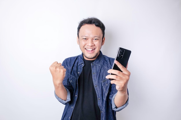 A young Asian man with a happy successful expression wearing navy blue shirt and holding smartphone isolated by white background