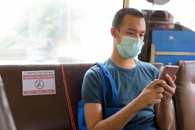 Young Asian man with mask using phone while sitting on the bus with distance