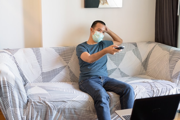 検疫の下で自宅でテレビを見てマスクを持つ若いアジア男