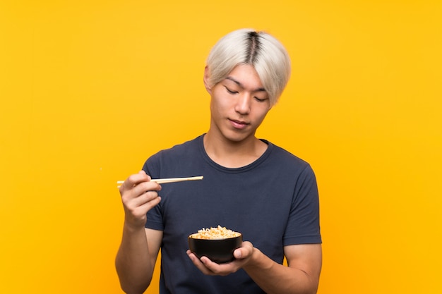 Young asian man with noodles over isolated yellow 