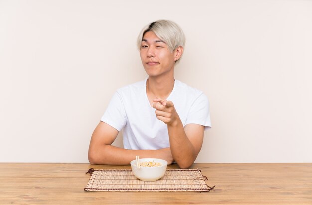 Young asian man with ramen in a table points finger at you