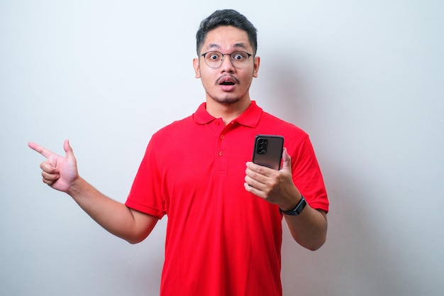 Young Asian Man with shock face holding a mobile phone pointing at copy space