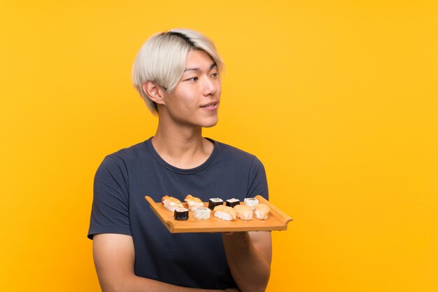 Young asian man with sushi over isolated yellow background looking to the side
